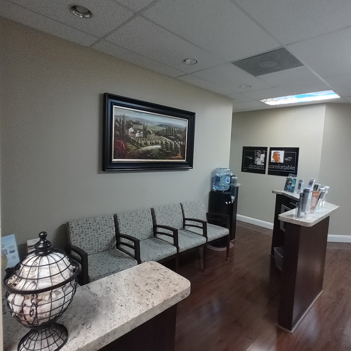 The image shows an interior view of a waiting area within a medical office, featuring a framed landscape painting on the wall, a reception desk with a lamp, a water fountain, a bench, and a potted plant.