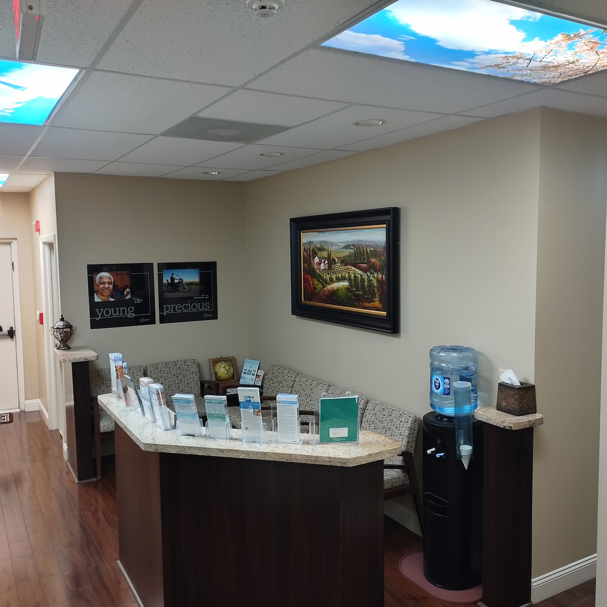 The image depicts a well-lit office waiting area with a reception desk, framed artwork on the wall, a water bottle dispenser, and various items on the countertop including brochures and a plant.