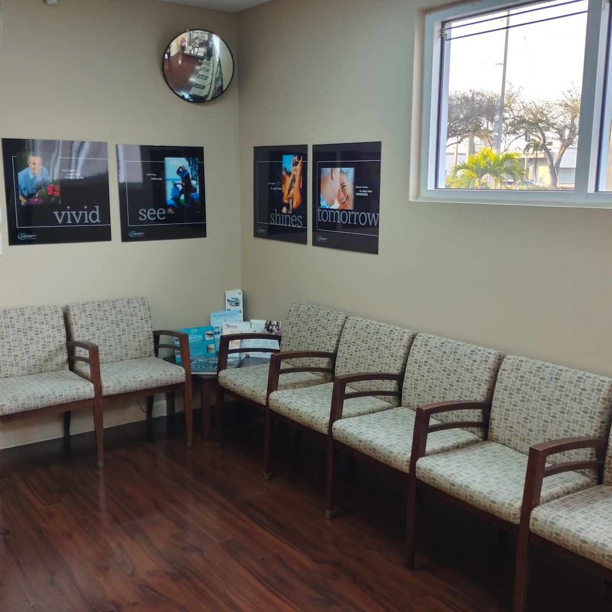 The image shows an interior space with a waiting area featuring a beige sofa, multiple chairs, a coffee table with printed materials, framed pictures on the wall, a window, and a clock.