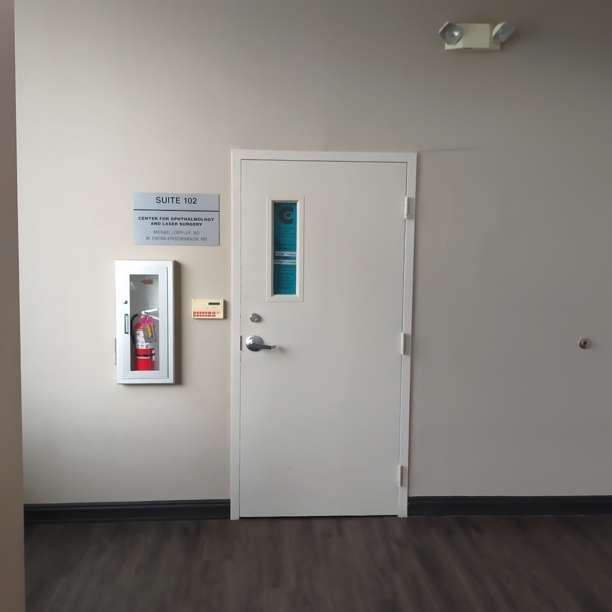 The image shows an interior view of a room with a closed door, featuring a sign indicating  Suite 12  above the door, a red fire extinguisher mounted on the wall next to the door, and a white emergency call box attached to the wall outside the door. The door has a window at the top, and there is a sign on the glass that reads  DO NOT ENTER.  The floor appears to be carpeted, and the walls are painted in neutral tones. There is no visible text providing information about the location or context of the image.