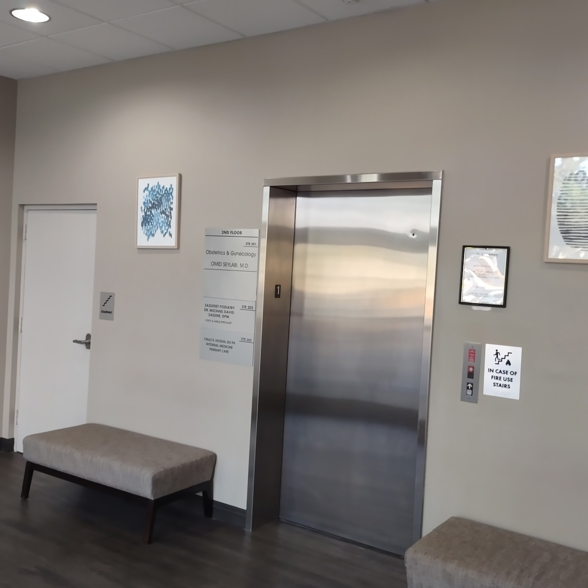 The image depicts an elevator lobby with a bench, framed artwork on the wall, a door with a sign, and a security card reader.