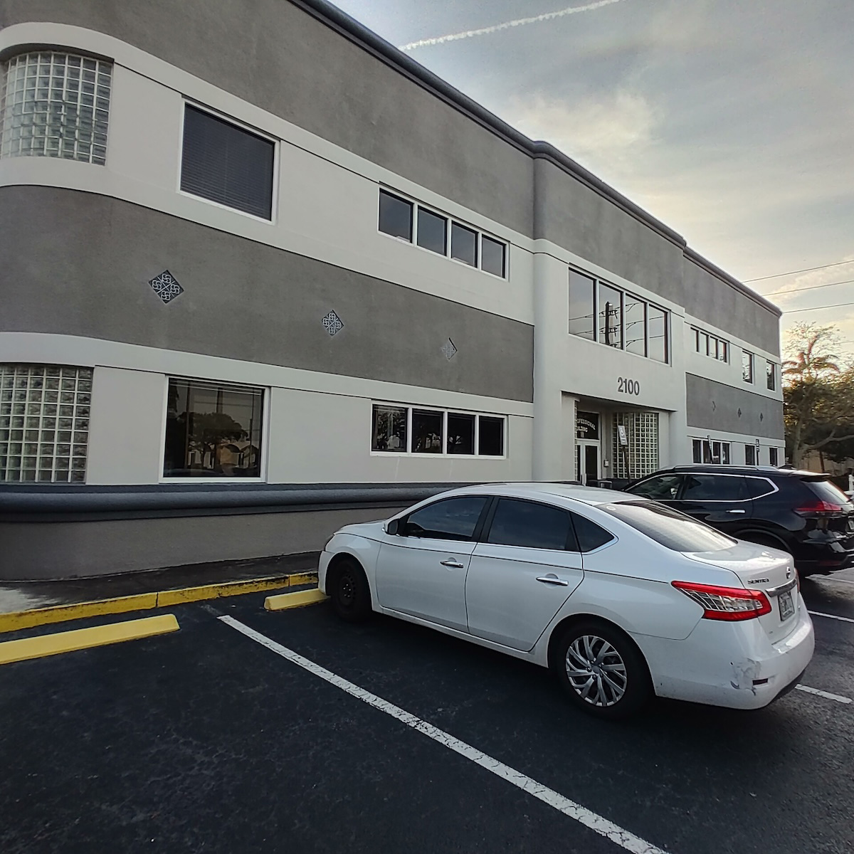 The image shows a white car parked in front of a large, modern building with a flat roof, featuring a glass facade with multiple windows and a prominent entrance.