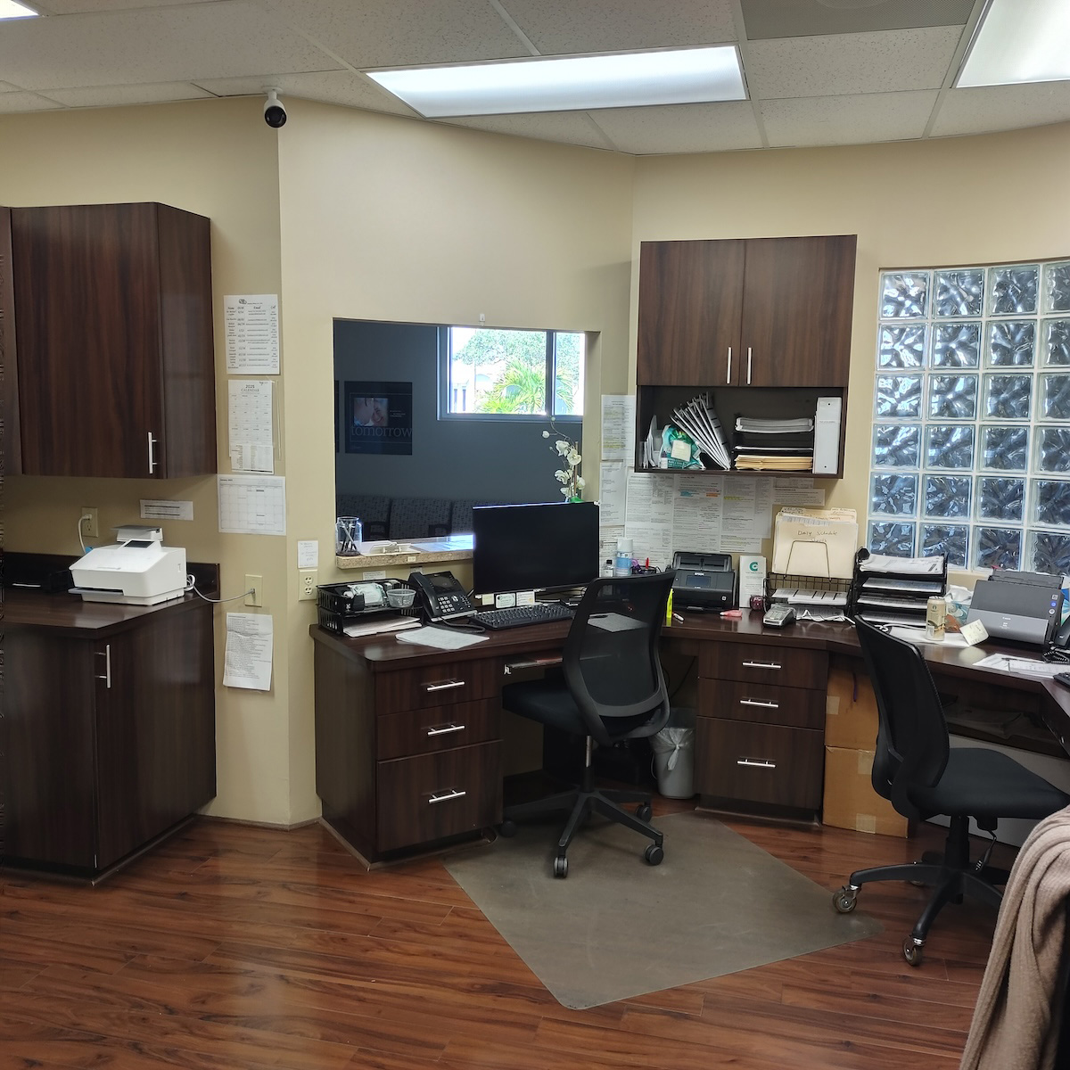 The image shows an office space with wood flooring, a desk area with a computer monitor, keyboard, and mouse, a receptionist s counter with a window behind it, a waiting area with chairs, a cabinet with a mirror above it, and various office equipment.