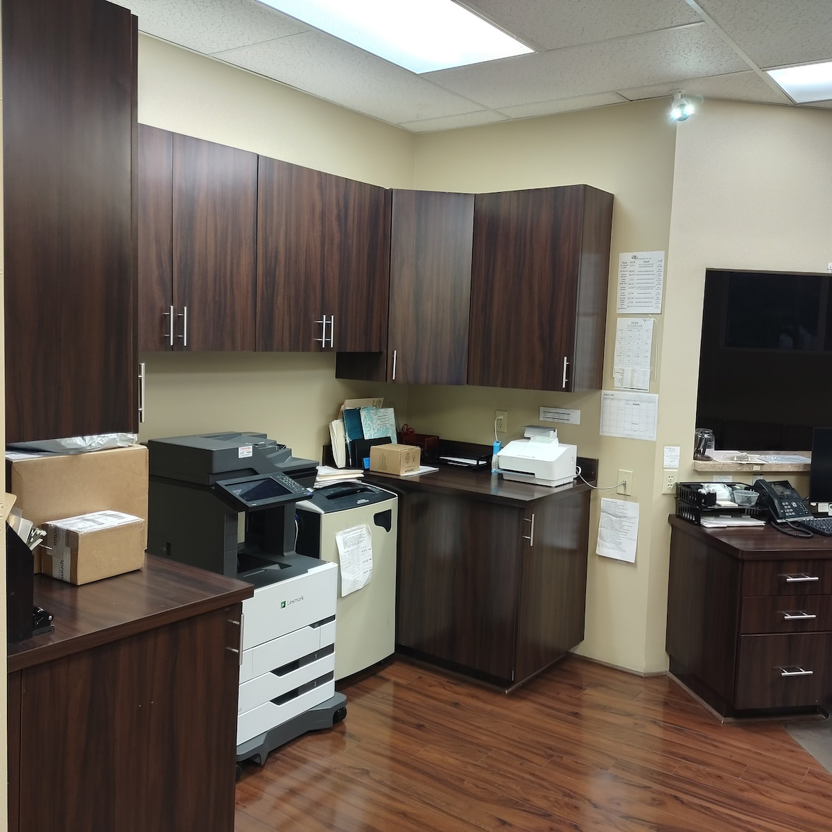 The image shows an office space with a printer, scanner, and computer on a desk, along with cabinets and a counter area with a keyboard and monitor.