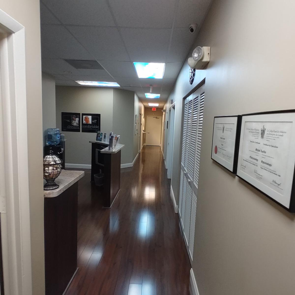 The image shows an interior hallway within a building, featuring polished flooring, white walls, and a series of framed certificates on the right wall.