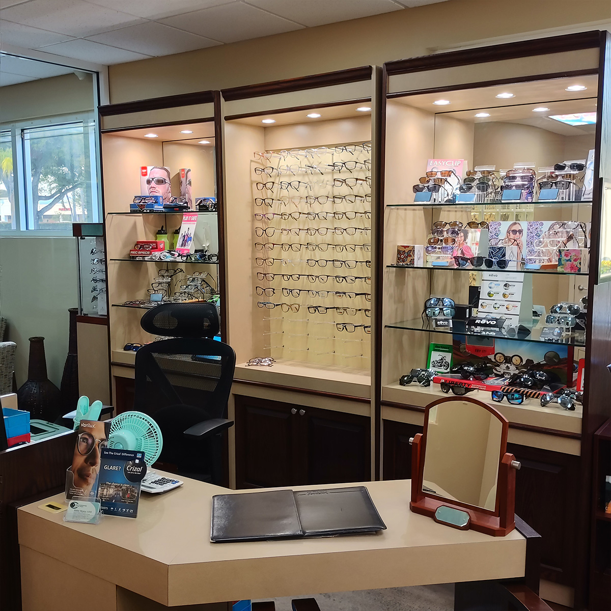 The image shows an optical store display with various frames on shelves behind a glass wall and a desk in front with a chair, indicating a retail setting for eyewear sales.