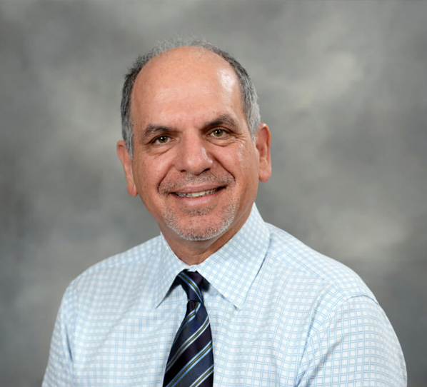 The image shows a man smiling at the camera with his mouth open, wearing a white shirt with a blue tie, dark pants, and glasses. He has short gray hair, and the background suggests an indoor setting with a wall-mounted light fixture and a checkered pattern on the wall.