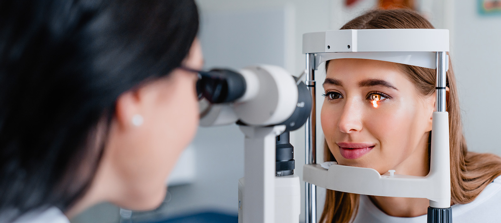 A woman with glasses and an eye patch sits at an optometrist s station while another person, possibly a technician, looks into her eye with a device.