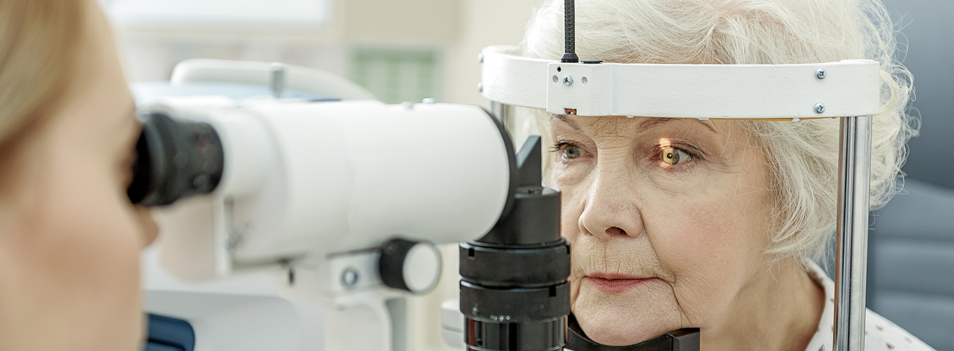 The image depicts an elderly woman seated at an eye examination machine with a technician standing beside her.