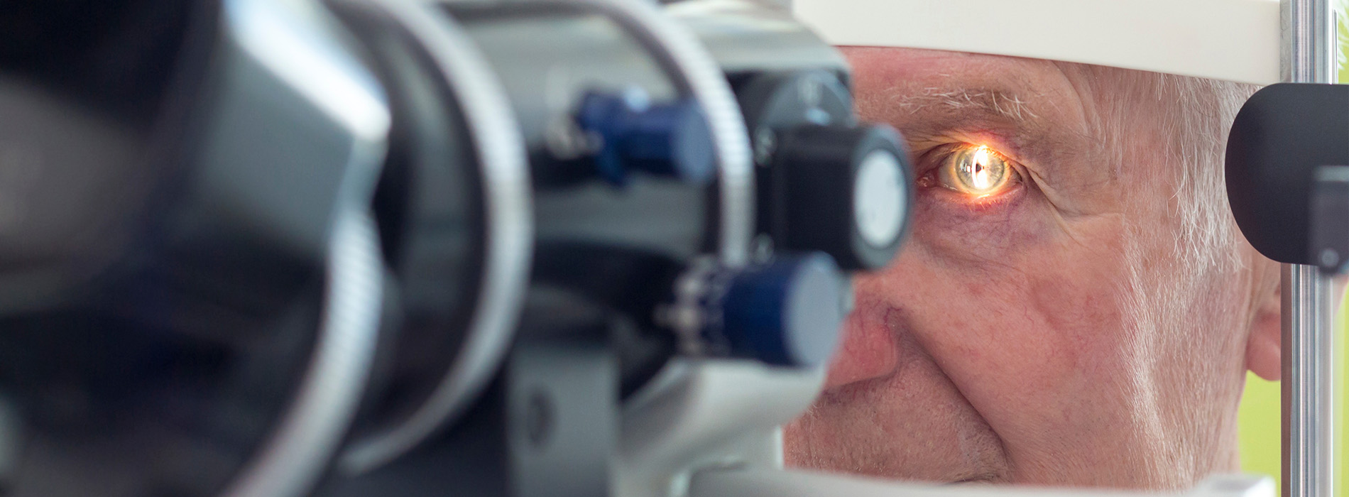 The image shows an elderly man sitting at an eye examination machine with his eyes open, looking towards the camera, while a device with a blue light illuminates his eye.