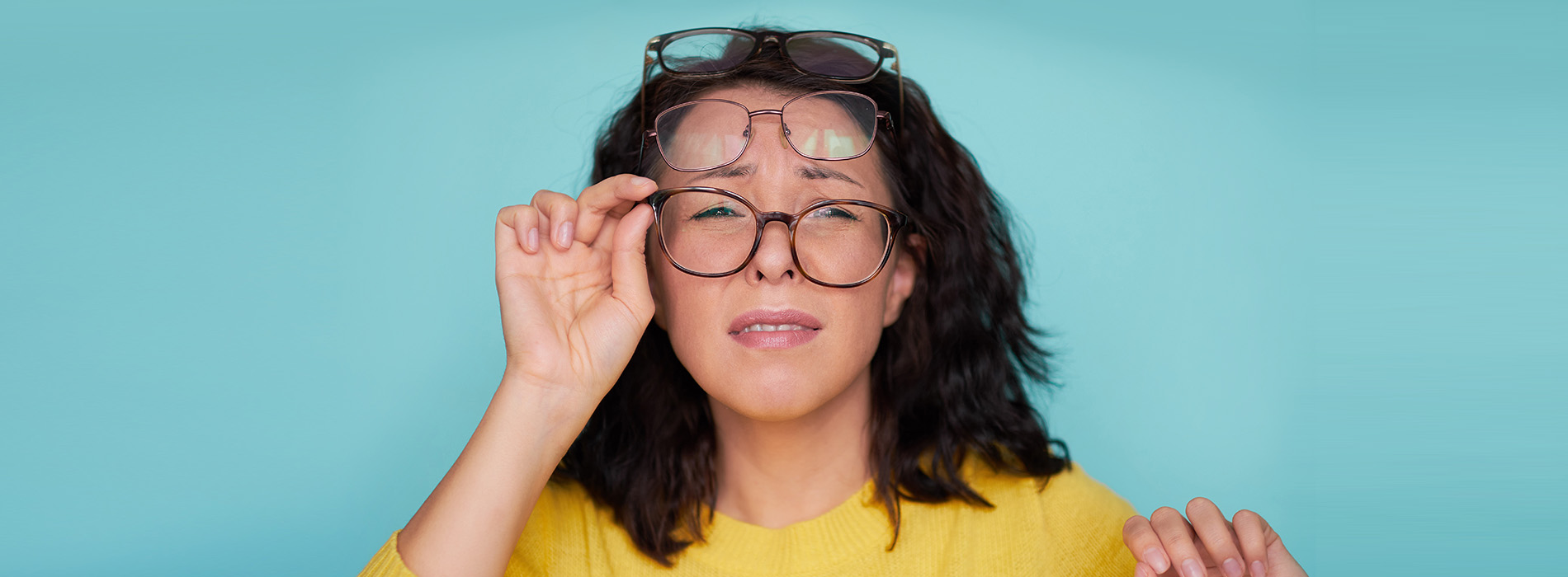 A woman wearing glasses holds up her hand, with a blurred background in shades of blue and green.