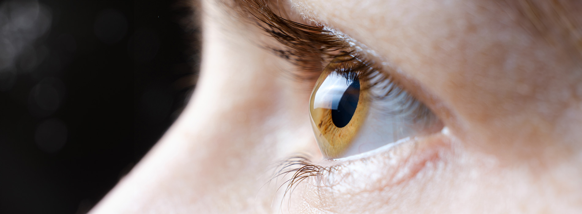 The image shows a close-up of a human eye with a reflection of light on its surface, set against a blurred background that appears to be a face.