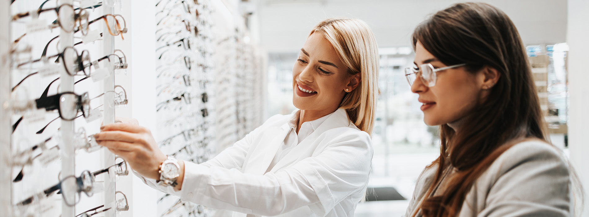 Two women shopping at an optical store, with one woman reaching out to touch a pair of glasses on display.