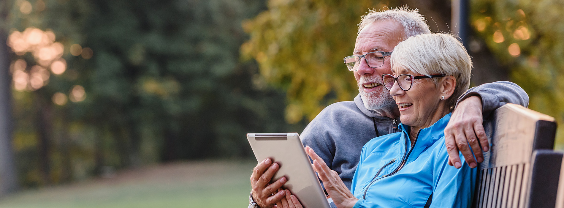 An elderly couple sitting on a bench, sharing a moment together while looking at something on a tablet.