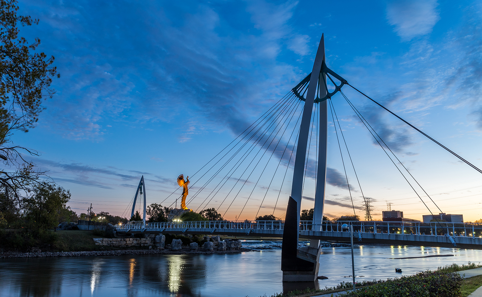 The image shows a scenic view at dusk with a bridge over a river, illuminated by streetlights, with a city skyline in the background and a clear sky above.