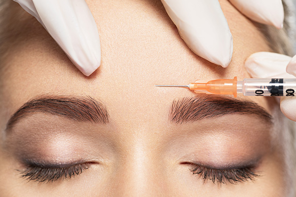 This is an image of a person receiving cosmetic treatment, with a syringe being injected into their skin near the eye area, while they are wearing makeup and lying down under medical supervision.