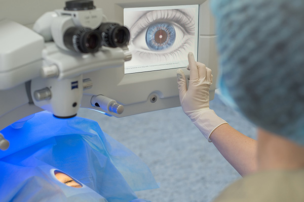 A person wearing medical protective gear is operating an ophthalmological machine while examining a patient s eye, with a large screen displaying the eye s details.