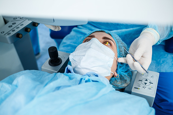 A medical professional wearing a surgical mask and blue gown is performing a procedure on a patient lying on an operating table, with a monitor displaying vital signs above them.