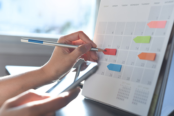 An individual s hand holding a pen is seen writing on a digital calendar displaying a month s schedule with color-coded events.