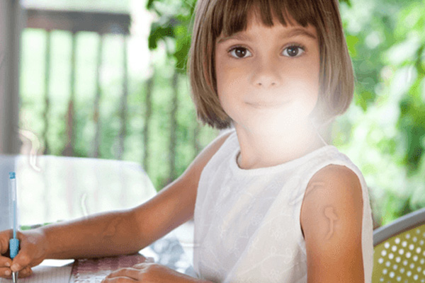 A young girl sitting at a desk with a notebook, pen, and a laptop, smiling and looking towards something outside the frame.