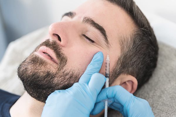 The image features a man lying down with his eyes closed while receiving a medical treatment, possibly a facial procedure, with a syringe being injected into his skin by someone out of frame.