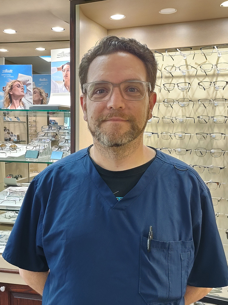 The image shows a man standing in front of an optical store display case, wearing glasses and a stethoscope badge, suggesting he is an optician with medical training.