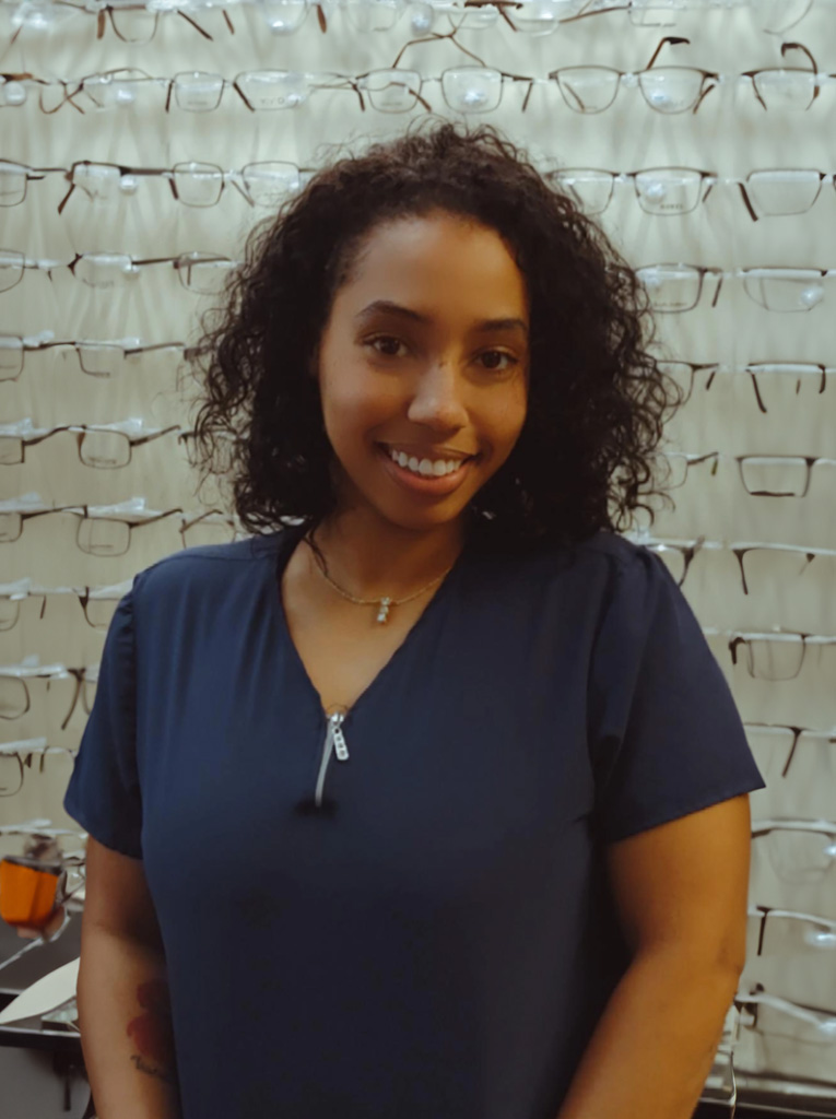 The image features a person standing in front of an array of eyeglass frames, smiling at the camera.