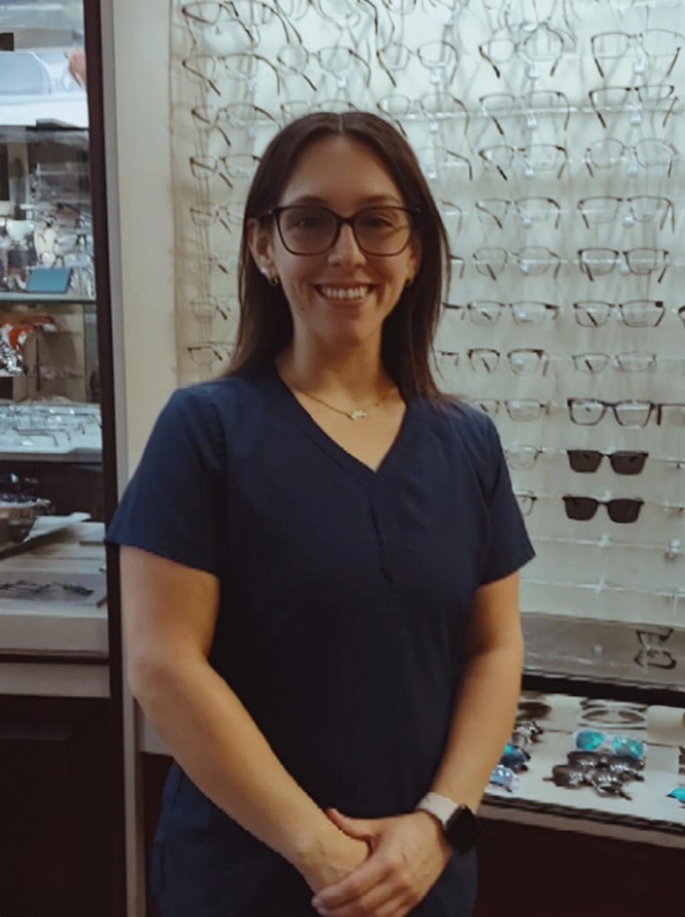 The image shows a woman standing behind a display case filled with eyeglasses, wearing glasses herself, and smiling at the camera.