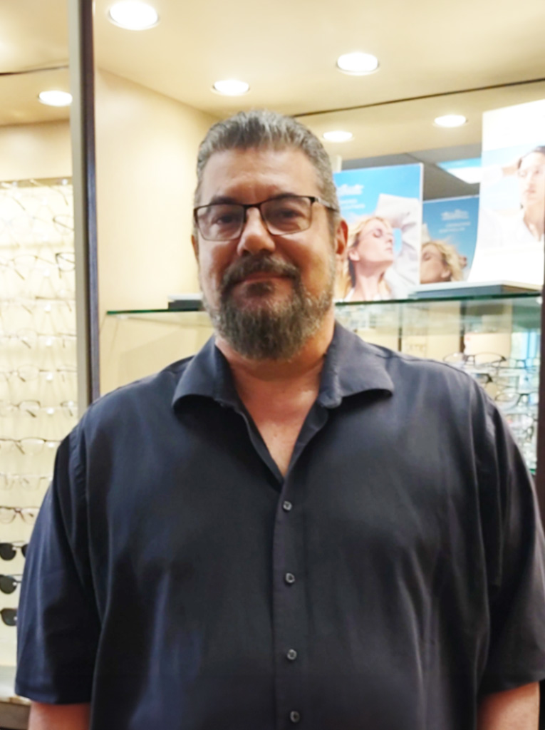 The image shows a man standing inside an optician s store, wearing glasses and a black shirt, with a beard and mustache, smiling slightly at the camera.