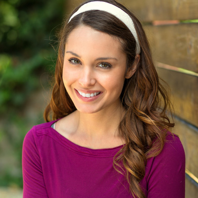 A woman with long brown hair smiling at the camera wearing a purple top and a white headband.