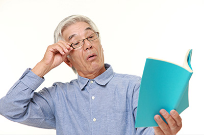 An elderly man with glasses holding an open book, looking upwards with his hand on his forehead.