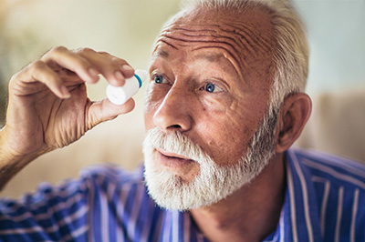 The image depicts an elderly man with a mustache and beard, holding a magnifying glass up to his eye, examining something closely, while seated indoors.