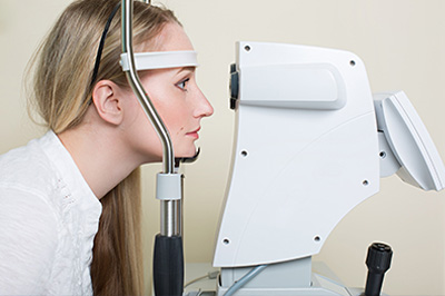 An adult female with blonde hair wearing a white lab coat and a headset is seated in front of an optical device, possibly a vision test machine, with her face close to the screen.