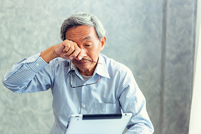 An elderly man with glasses, wearing a blue shirt, is seated at a desk with his head resting on his hand, looking down and away from the camera, while holding a tablet computer.