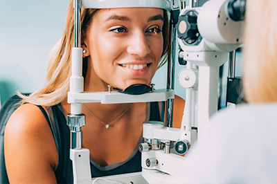 A woman sitting at an eye exam station, looking through a device while a technician checks her eyes with a machine.